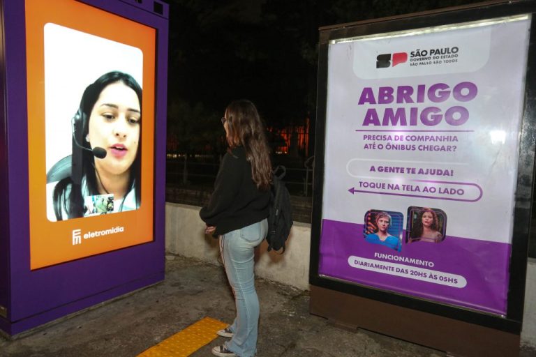 Mulher em pé conversa com uma atendente através de um painel do Abrigo Amigo instalado em um pontos de ônibus