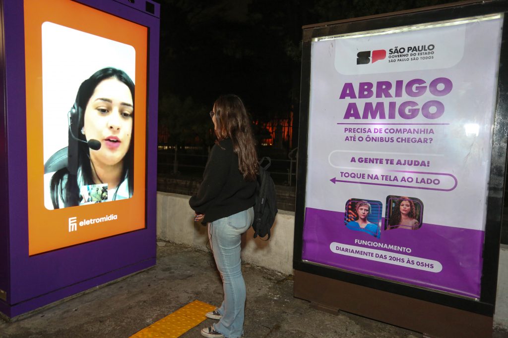 Mulher em pé conversa com uma atendente através de um painel do Abrigo Amigo instalado em um pontos de ônibus
