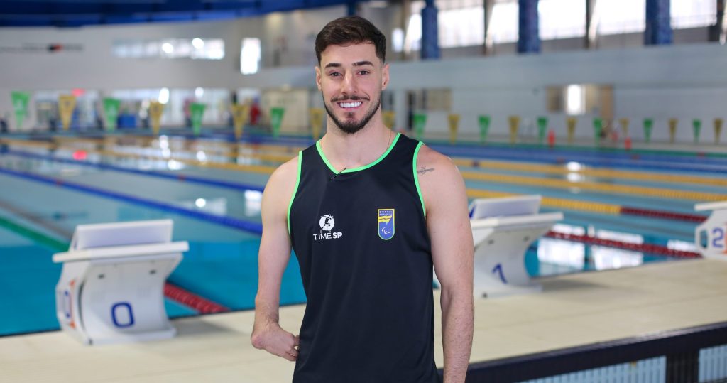 Lucas Mozela, nadador que estará nas Paralimpíadas 2024, aparece sorrindo em frente a uma piscina. Ele é um homem branco, de cabelos curtos, e usa uma camiseta regata característica do Time São Paulo