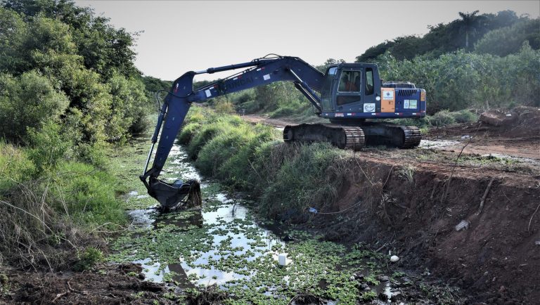 Retroescavadeira posicionada ao lado de um córrego retira resíduos da água. A paisagem conta com diversas árvores