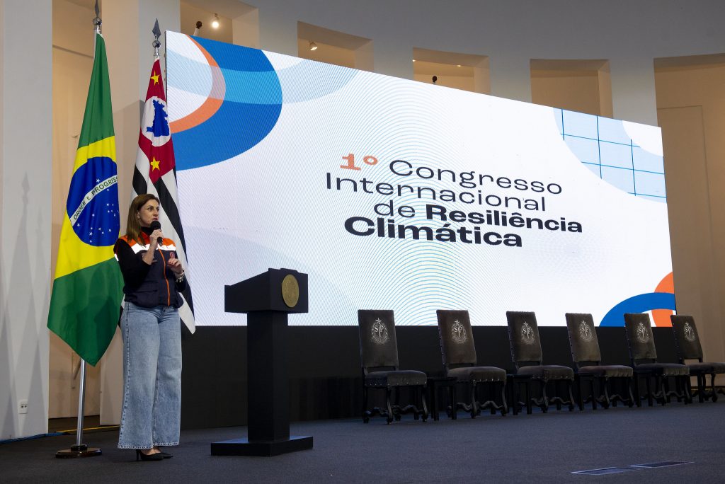 Cabo Zimmerman, uma mulher branca de cabelos ruivos, segura um microfone enquanto fala com um painel eletrônico do Congresso de Resiliência Climática ao fundo
