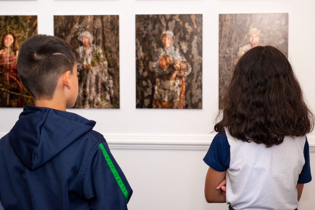 Duas crianças, um menino e uma menina, olhando para uma parede, de costas, com três quadros. As criançasusam uniformes escolares. A exposição se dá no Palácio Boa Vista